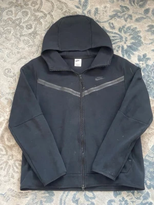 Svart Nike hoodie med dragkedja - [FRI FRAKT 🚚]Säljer en svart hoodie från Nike med hel dragkedja och huva. Den har diskreta gråa detaljer över bröstet och Nike-loggan på vänster sida. Materialet är mjukt och bekvämt, perfekt för en avslappnad look.