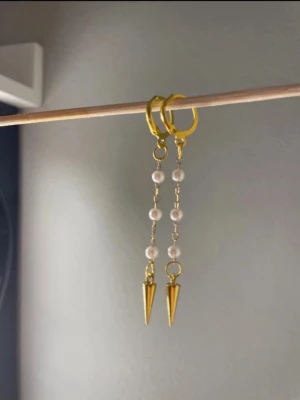 Guldiga örhängen med pärlor och spikes - Snygga örhängen i guldton med små vita pärlor på rad och avslutande guldfärgade spikes. Hängande modell med modern och edgy känsla, perfekta för att lyfta din stil. Materialet ser ut att vara metall och pärlor. Egengjorda 