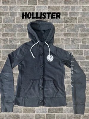 Mörkgrå hoodie/cardigan från Hollister - Snygg mörkgrå hoodie från Hollister med knäppning framtill, vita dragsnören i huvan och tryckt logga på bröstet. Hollister-text längs ena ärmen och två fickor framtill. Perfekt för en avslappnad och trendig look. Tröjan är i storlek M, men skulle säga att den har en väldigt liten passform. Skulle mer säga att den är i storlek S/XS. Om ni har några mer funderingar är det bara att fråga😊