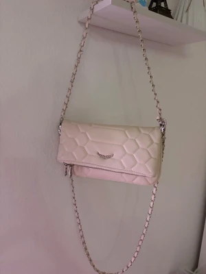 Beige quiltad axelväska med kedja - Snygg beige axelväska med quiltat mönster och silverfärgad kedjerem. Väskan har ett litet metallmärke framtill och dragkedja nedtill. Perfekt storlek för det viktigaste och ger en trendig touch till din outfit.