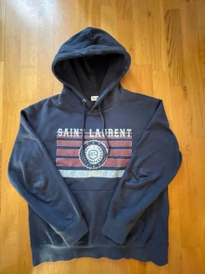 Mörkblå Saint Laurent hoodie - Snygg mörkblå hoodie från Saint Laurent med tryck framtill i vitt och rött. Perfekt för dig som vill ha en stilren och trendig look! Tröjan är i storlek M, bara att höra av er vid frågor! 😊