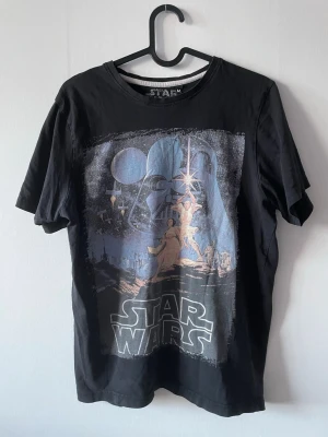 Svart Star Wars t-shirt - Svart t-shirt med Star Wars-tryck. Ett stort tryck framtill som visar Darth Vader och klassiska karaktärer från filmen. T-shirten har rund hals och korta ärmar. 