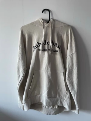 Beige hoodie Club de Basket Iowa - Snygg beige hoodie med svart broderad text 'club de basket' och 'University of Iowa' på bröstet. Klassisk känguruficka framtill och justerbar huva med snören. 