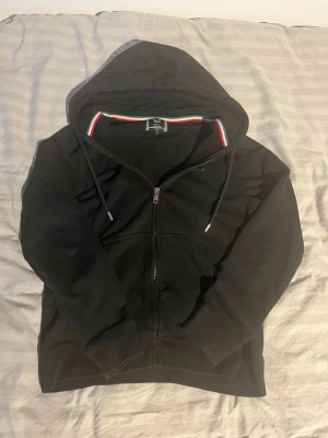 Svart vers@ce äkta hoodie - Säljer en svart hoodie med dragkedja. Hoodien har huva med dragsko, ribbade muddar och fickor framtill. Insidan av kragen har en snygg detalj med röd, vit och grön rand. Perfekt för en chill och avslappnad stil. Köpt på johnells