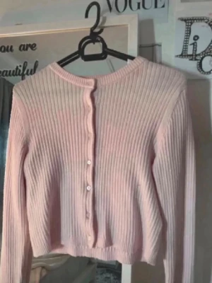 Ljusrosa ribbad kofta med knappar - Söt ljusrosa kofta med ribbad struktur och rund halsringning. Koftan har långa ärmar och knäpps framtill med små vita knappar. Perfekt för lager på lager och ger en mjuk, feminin vibe.