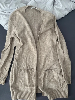 Beige lång kofta från Only - Mysig beige lång kofta från Only med två stora fickor framtill och ribbade muddar vid ärmslut och nederkant. Koftan är öppen framtill och har en avslappnad passform, perfekt att slänga över en t-shirt eller topp. Nopprig på sidan 