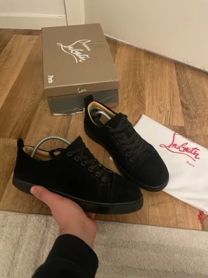 Svarta sneakers från Christian Louboutin - Säljer ett par svarta sneakers från Christian Louboutin i mocka med matchande svarta skosnören och sula. Klassisk låg modell med diskret design och exklusiv känsla. Kommer med originalkartong och dustbag. Obs ett litet hål finns vid hälen 