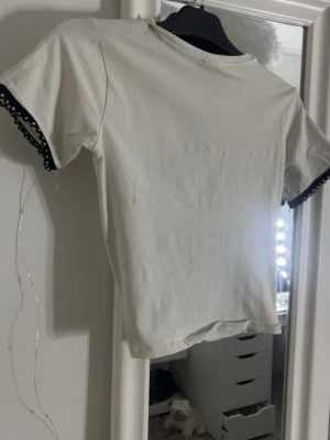 Vit topp med svarta pärldetaljer - Snygg vit kortärmad med  svarta kanter dekorerade med små pärlor. T-shirten har en enkel, rak passform och är tillverkad i mjukt bomullstyg. Perfekt för dig som gillar unika detaljer på klassiska plagg.