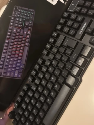 FOREV svart gamingtangentbord - Säljer ett svart gamingtangentbord från FOREV med klassisk layout och tydliga vita bokstäver på tangenterna. Tangentbordet har en robust plastdesign och RGB-belysning som syns på kartongen. Perfekt för gaming eller datorarbete.