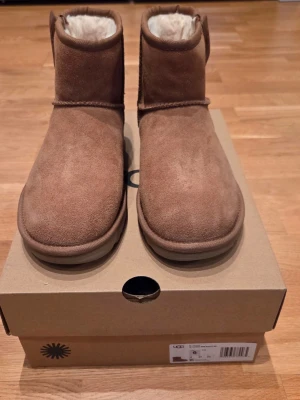 Bruna UGG Classic Mini stövletter - Bruna UGG Classic Mini stövletter i mocka med mjukt, vitt foder på insidan. Klassisk rund tå och låg skaftlängd. Sitter på en robust, räfflad sula för extra grepp. Perfekta för dig som vill ha både stil och komfort under kalla dagar.