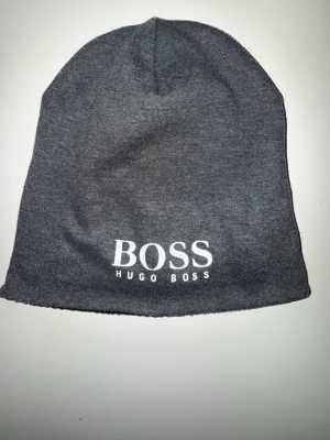 Grå mössa från Hugo Boss - Snygg grå mössa från Hugo Boss med vit logga framtill. Tillverkad i mjukt och stretchigt material som sitter skönt på huvudet. Perfekt accessoar för att hålla värmen och addera stil till din outfit.