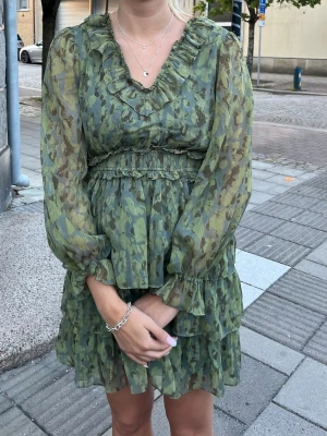 Grön volangklänning från Aware - Hej!☀️Säljer en grön kortklänning från Aware med volanger och v-ringning. Klänningen har långa ärmar och ett diskret blommigt mönster. Resår i midjan och volangdetaljer både vid halsringning och kjol. Väldigt fin klänning. Använd vid ca 2 tillfällen💕
