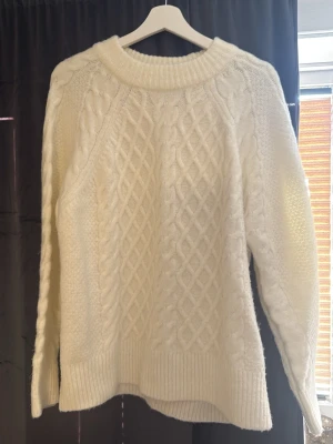 Vit kabelstickad tröja - Säljer en krämvit kabelstickad tröja med grov struktur och ribbade muddar. Tröjan har rund halsringning och långa ärmar. Perfekt för kalla dagar och ger en mysig, trendig look. Lappen med storlek är avklippt men är i storlek S