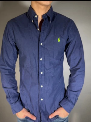 Marinblå skjorta från Ralph Lauren - Klassisk marinblå skjorta från Ralph Lauren med button-down krage och vita knappar. Ikoniska gröna Polo-loggan broderad på bröstet. Skjortan är långärmad och tillverkad i mjuk bomull, perfekt för en clean och stilren look.