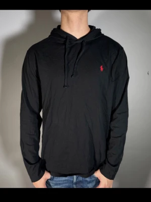 Svart hoodie från Ralph Lauren - Svart hoodie från Ralph Lauren med klassisk röd logga broderad på bröstet. Hoodien har dragsko i huvan och är tillverkad i mjukt bomullstyg. Perfekt för en chill och stilren look.