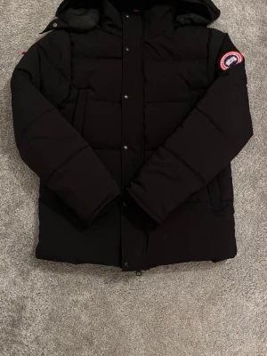 Svart dunjacka från Canada Goose - Svart dunjacka från Canada Goose med huva och klassisk logotyp på ärmen. Jackan har raka, quiltade paneler, stora fickor och stängs med både dragkedja och knappar. Perfekt för kalla vinterdagar och riktigt varm.