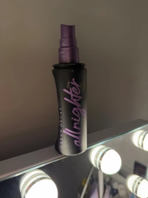 Urban Decay Setting spray - Urban Decay All Nighter face mist i svart plastflaska med lila spraymunstycke och text. Perfekt för att fixera makeup och ge en fräsch känsla hela dagen. Flaskan har en smidig och modern design med tydlig logotyp. Flaskan är full upp till bokstaven E 