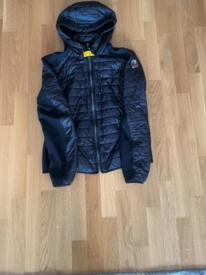 Svart quiltad jacka med huva Parajumpers - Snygg svart quiltad jacka från Parajumpers med huva och dragkedja framtill. Jackan har ett emblem på ärmen och detaljer i mesh på sidorna och ärmarna. Perfekt för dig som vill ha en sportig och stilren look.