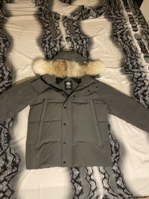 Grå dunjacka med pälskantad huva - Snygg grå dunjacka från Canada Goose med stor huva och avtagbar pälskant i beige och brunt. Jackan har flera praktiska fickor framtill, dragkedja och knappar. Perfekt för kalla vinterdagar och riktigt varm tack vare dunfyllningen.