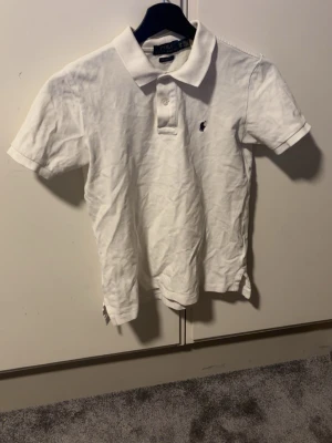 Vit pikétröja från Polo Ralph Lauren - Vit pikétröja från Polo Ralph Lauren med klassisk krage, korta ärmar och två knappar framtill. Tröjan har en liten broderad logga på bröstet och är gjord i mjuk bomull för en clean och stilren look.