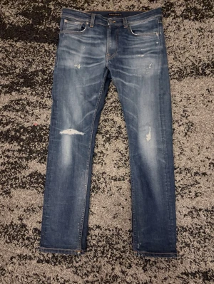 Blå slitna jeans från Nudie Jeans - Säljer ett par blå jeans från Nudie Jeans med skinny fit och slitningar på  framsidan. Jeansen har klassiska fem fickor, orange sömmar och är gjorda i mjuk denim. Perfekta för en avslappnad och trendig look.