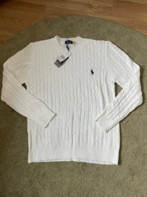 Vit kabelstickad tröja Polo Ralph Lauren - Kabelstickad vit tröja från Polo Ralph Lauren med rund halsringning och den klassiska mörkblå logotypen broderad på bröstet. Tröjan har långa ärmar och ribbade muddar vid ärmslut och nederkant. Perfekt för en clean och stilren look. Den är aldrig andvänd och i storleken s men skulle säga att den är mer som en storlek m