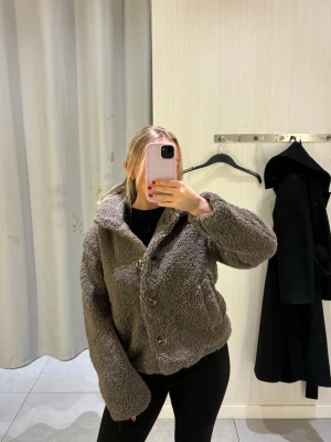 Ny oanvänd Brun teddyjacka med stora knappar - Mysig brun teddyjacka med stora, synliga knappar framtill och bred krage. Jackan har en boxy passform och är gjord i fluffigt, teddyliknande material som ger en trendig sopranovibe. Helt ny och oanvänd ursprungligt pris 550kr