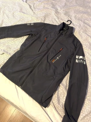 Mörkgrå vindjacka från Sail Racing - Snygg mörkgrå vindjacka från Sail Racing med dragkedja framtill, två bröstfickor med orange detaljer och logga på bröstet samt ärmen. Jackan har hög krage och är tillverkad i ett lätt, vindtätt material som passar perfekt för blåsiga dagar.