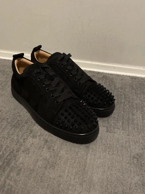 Svarta Louboutin sneakers med nitar - Säljer ett par svarta sneakers från Christian Louboutin med mattsvarta nitar på tån och klassisk röd sula. Skorna har snörning, låg profil och är tillverkade i mocka och läder. Perfekta för dig som vill sticka ut med en edgy och lyxig stil.