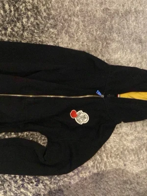 Svart hoodie från Moncler med dragkedja - Snygg svart hoodie från Moncler med guldfärgad dragkedja och stor logga på bröstet. Klassisk huva och fickor framtill. Insidan av huvan har gul detalj och tricolore-band syns vid dragkedjan. Perfekt för en chill och stilren look.