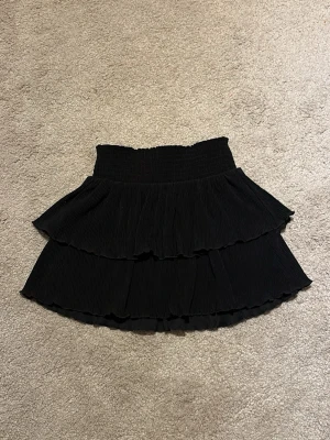 Svart volangkjol från Kappahl - Säljer en svart kortkjol från Kappahl med bred resår i midjan och dubbla volanger. Storlek 134-140 (dock mycket stretchig och kan nog passa större storlekar) säljer eftersom den inte används 🫶skriv om du har någon fråga!