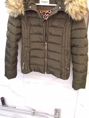 Olivgrön pufferjacka med fuskpäls - Snygg olivgrön pufferjacka från Rocco Barocco med avtagbar huva och fuskpäls. Jackan har två dragkedjefickor fram, quiltad design och leopardmönstrat innerfoder. Perfekt för kalla vinterdagar och riktigt trendig look.