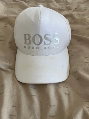 Vit keps från Hugo Boss - Snygg vit keps från Hugo Boss med stort silverfärgat BOSS-tryck framtill. Klassisk böjd skärm och justerbar baksida. Tillverkad i bomullsmix för en bekväm och luftig känsla. Perfekt accessoar för en clean och stilren look. Den har svett fläckar intill sig samt lite framtill men går säkert bort med klorin! Qr koden finns även intill kepsen 