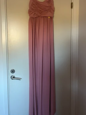 Lång rosa klänning med draperad topp - Säljer en elegant långklänning i mjukt rosa tyg med breda axelband och draperad överdel. Klänningen har ett flytande fall och markerad midja, perfekt för speciella tillfällen. Aldrig använd 