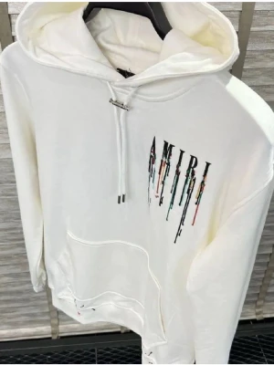 Amiri - Säljer en vit hoodie från Amiri med färgstarkt droppande logotyptryck på bröstet. Hoodien har huva med dragsko och en stor magficka. Materialet är mjukt och skönt, perfekt för en avslappnad streetwear-look.