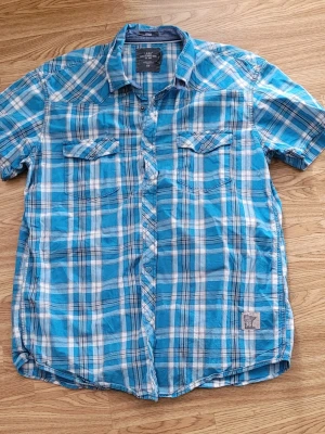 Blåvit rutig kortärmad skjorta  - Snygg blå och vit rutig skjorta från HM logg med korta ärmar och två bröstfickor med knappar. Skjortan har klassisk krage och knäppning framtill. Perfekt för en avslappnad stil och tillverkad i mjuk bomull.