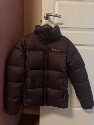 Brun pufferjacka från Columbia - Snygg brun pufferjacka från Columbia med hög krage och dragkedja framtill. Jackan har en quiltad design med breda paneler och diskret logga på bröstet. Perfekt för kalla vinterdagar och riktigt varm tack vare det tjocka materialet. På andra bilden ser man att det är några få fläckar ovanför loggan men inte något som läggs märke till