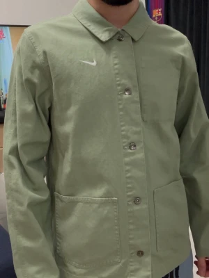 Grön overshirt jacka från Nike - Snygg ljusgrön overshirt jacka från Nike med klassisk krage och vita broderade loggan på bröstet. Jackan har knäppning framtill, tre fickor och är tillverkad i bomull. Perfekt för lager på lager och enkel att styla till olika outfits.