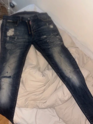 Dsquared2 Mörkblå slitna jeans med detaljer - Säljer ett par mörkblå jeans med slitningar och färgstänk för en cool, trendig look. Jeansen har normal passform och klassisk femficksdesign. Perfekta för dig som gillar streetstyle och vill sticka ut lite extra.