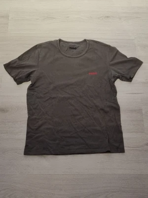 Mörkgrå t-shirt från Hugo - Snygg mörkgrå t-shirt från Hugo med diskret röd logga på bröstet. Klassisk rund halsringning och korta ärmar. Tillverkad i mjuk bomull som känns skön mot huden. Perfekt basplagg för en clean och enkel stil.