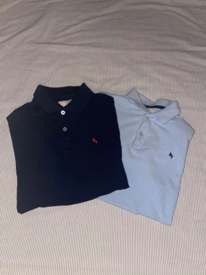 2-pack Abercrombie pikétröjor - Säljer två klassiska pikétröjor från Abercrombie & Fitch. En är marinblå med röd logga och den andra ljusblå med mörkblå logga. Båda har krage, knappar framtill och är gjorda i mjuk bomull. Perfekta till jeans eller shorts.