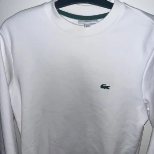 Vit sweatshirt från Lacoste - Klassisk vit sweatshirt från Lacoste med rund halsringning och det ikoniska gröna krokodilmärket broderat på bröstet. Tröjan har en relaxed passform och är tillverkad i mjukt bomullsmaterial. Perfekt för en clean och stilren look.