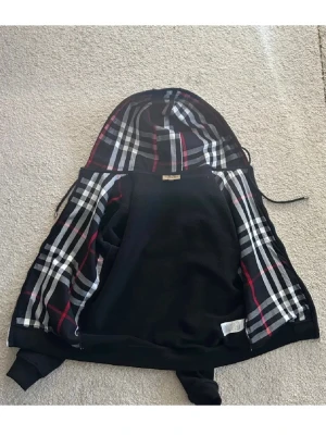 Burberry hoodie - Iprincip helt ny, använd 1-2 gånger!      Snygg svart hoodie från Burberry med dragkedja och klassiskt rutigt foder i huvan och insidan. Utsidan är helsvart med ribbade muddar och fickor framtill. Huvan har dragsko och insidan har det ikoniska Burberry-mönstret i svart, vitt och rött.