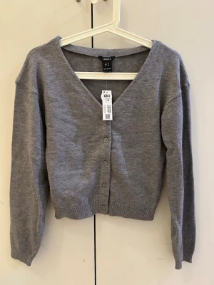  NyGrå kofta från Lindex med knappar - Snygg grå kofta från Lindex med v-ringning och knäppning framtill. Tillverkad i mjukt material och har ribbade muddar vid ärmslut och nederkant. Perfekt att slänga över en t-shirt eller linne för en chill look.