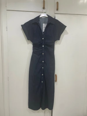 Denim klänning från zara - Snygg mörkblå klänning med v-ringning, markerad midja och knappar längs hela framsidan. Klänningen har korta ärmar och en rak, något längre passform. Perfekt för dig som gillar enkel men stilren design. Aldrig haft på mig den, fortfarande prislappen på, Storlek M men är som S eftersom den är liten i storlek, ord pris 500 och helt slut i lager