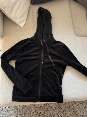 Svart velourhoodie från Gina Tricot - Mjuk och skön svart hoodie i velour från Gina Tricot. Modellen har dragkedja framtill, stor huva med snören och en praktisk magficka. Perfekt för en chill och avslappnad stil. Materialet är lent och ger en lyxig känsla.