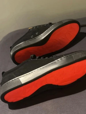 Svarta sneakers med röd sula - Snygga svarta sneakers med klassisk låg profil och kontrasterande röd yttersula. Skorna har svarta skosnören, metallöljetter och en diskret svart ovandel i textil och syntet. Insidan är beige för extra stilpoäng. Perfekta för dig som vill sticka ut med detaljer.