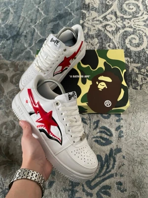 BAPE STA vita sneakers med röd stjärna - [FRI FRAKT 🚚]Snygga vita BAPE STA sneakers med röd stjärna på sidan och hajtandsdetalj i svart och vitt. Klassisk siluett med perforerad tå, vita skosnören och märkning på plösen. Kommer med originalkartong i camomönster. Perfekta för dig som gillar streetwear och unika detaljer.