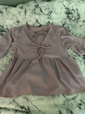 Ljusrosa kortklänning med knyt - Supersöt ljusrosa kortklänning med trekvartsärm och två dekorativa knyt framtill. Klänningen har en lätt rynkad kjoldel och är gjord i ett mjukt, ribbat material som ger en avslappnad men ändå stylish look.