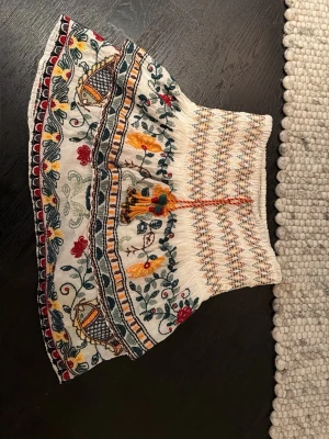 Broderad kortkjol med volang - Unik kortkjol i vitt med färgglad brodyr i rött, gult, grönt och blått. Kjolen har volangkant, smock i midjan och dekorativa tofsar framtill. Mönstret är blommigt och detaljerat, vilket ger en bohemisk vibe. Perfekt för dig som gillar statement-plagg! Den är lite insydd i middan men går lätt att sprätta upp